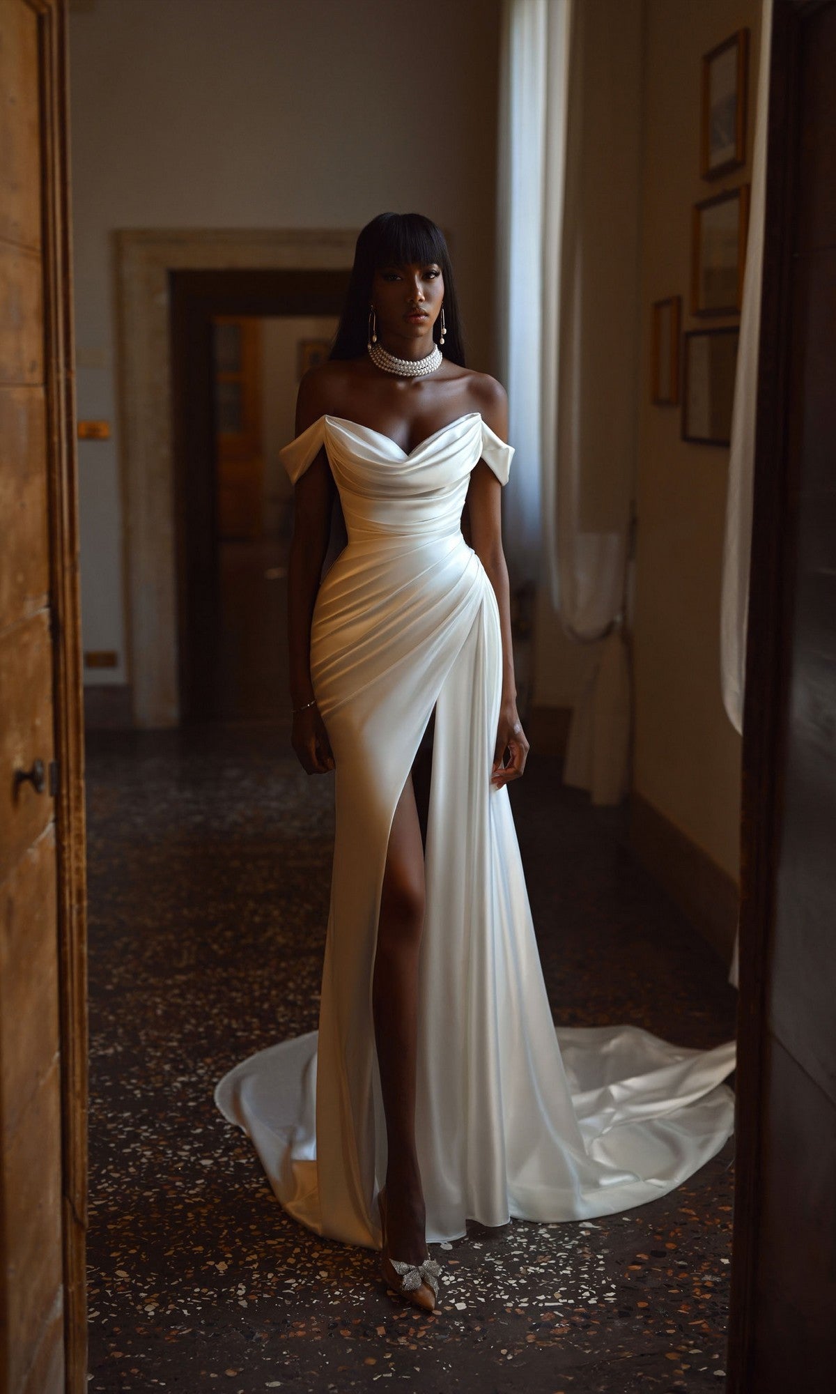A woman in the Alyce 7106 Long White Dress, featuring a cowl neckline and thigh-high slit, stands in a softly lit room with stone floors and framed pictures on the wall.